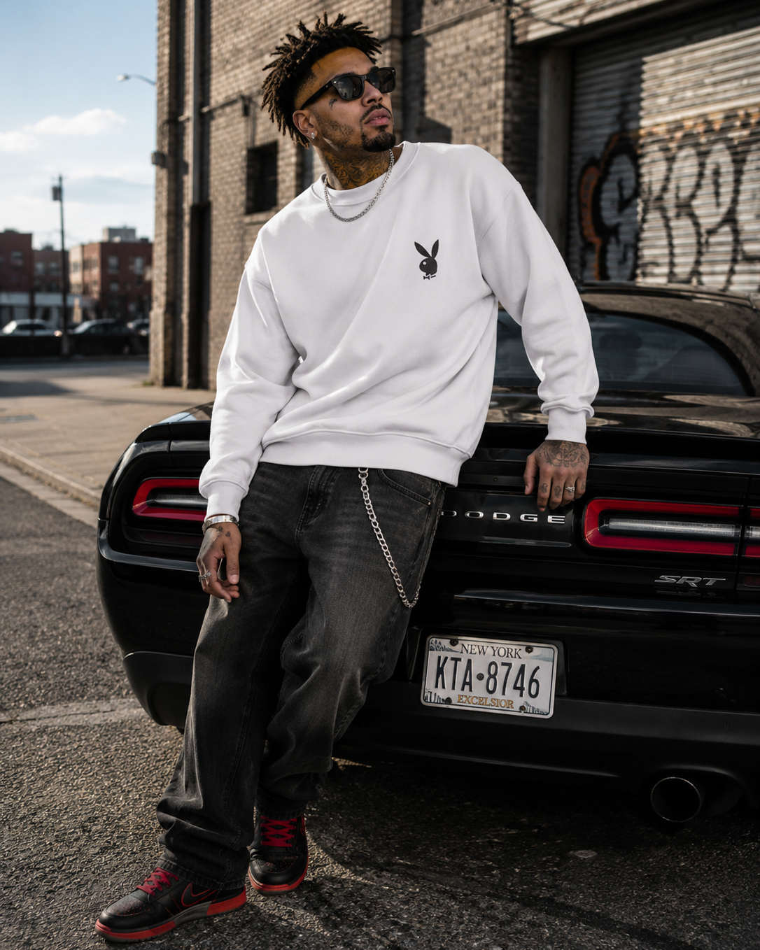 A man wearing a white Playboy sweatshirt, dark gray jeans, and red-and-black sneakers leans against a black Dodge SRT car in an urban street setting. #Streetwearmen