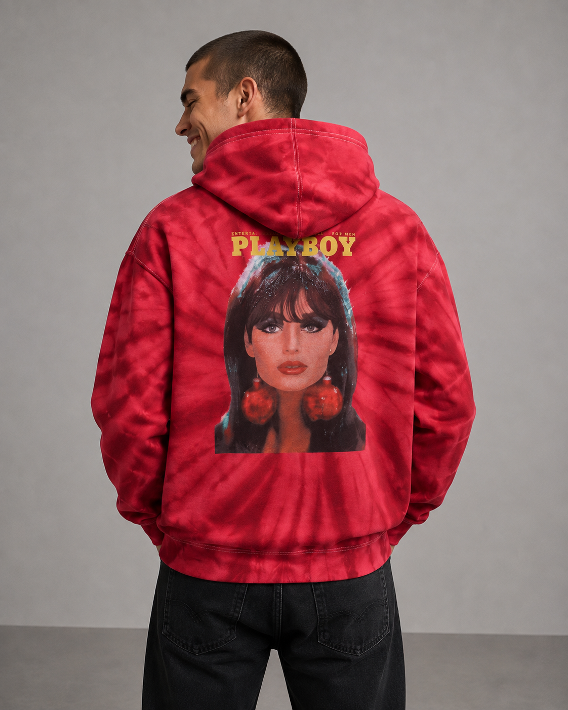 A man wearing a red tie-dye hoodie with a large graphic print of a woman's face and the word 'PLAYBOY' on the back, shown from behind against a gray studio background.