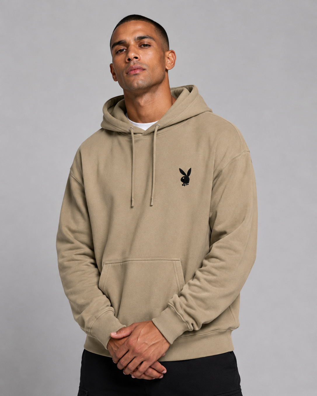 A man wearing a beige pullover hoodie with a black embroidered Playboy bunny logo on the left chest, paired with black cargo pants, against a solid gray studio background.