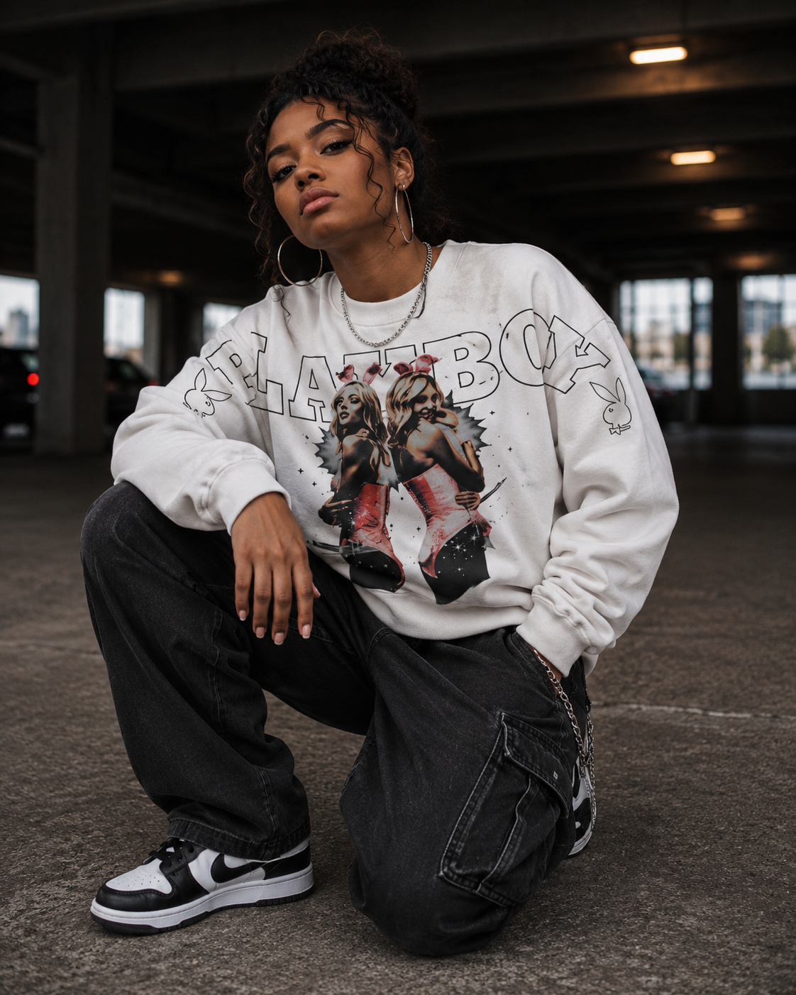 A person crouching in a concrete parking structure wearing a white graphic sweatshirt with a large illustrated print and outlined text, black cargo pants, and black-and-white sneakers. #Streetwearwomen