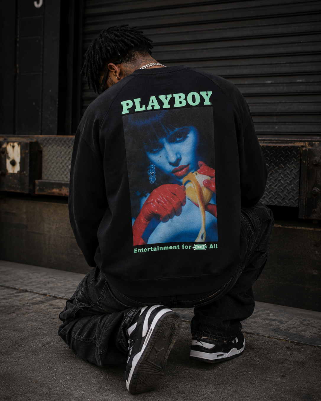 A person kneeling on a concrete street in front of a closed metal garage door, viewed from behind. They are wearing a black sweatshirt with a large back graphic featuring the word PLAYBOY in green and a stylized portrait with red gloves and a yellow object.  #Streetwearmen