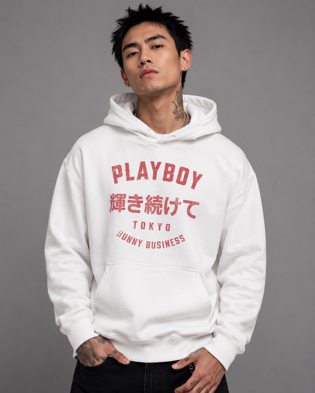 A man wearing a white Playboy hoodie with red text on the front, paired with black pants, standing against a solid gray studio background.