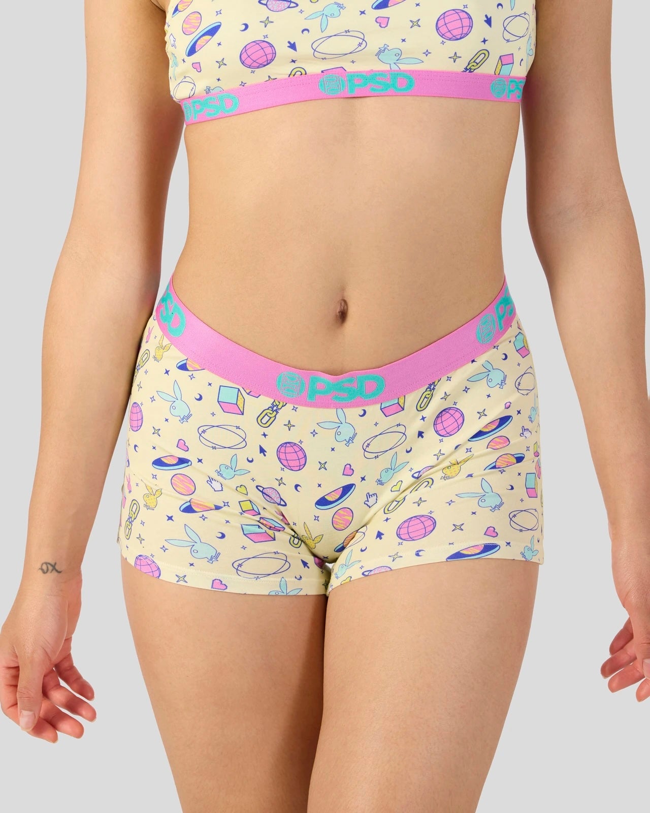 A model wearing a two-piece underwear set with a pastel multicolor space-themed print on a cream background, featuring a pink waistband with the brand name 'psd' in teal.