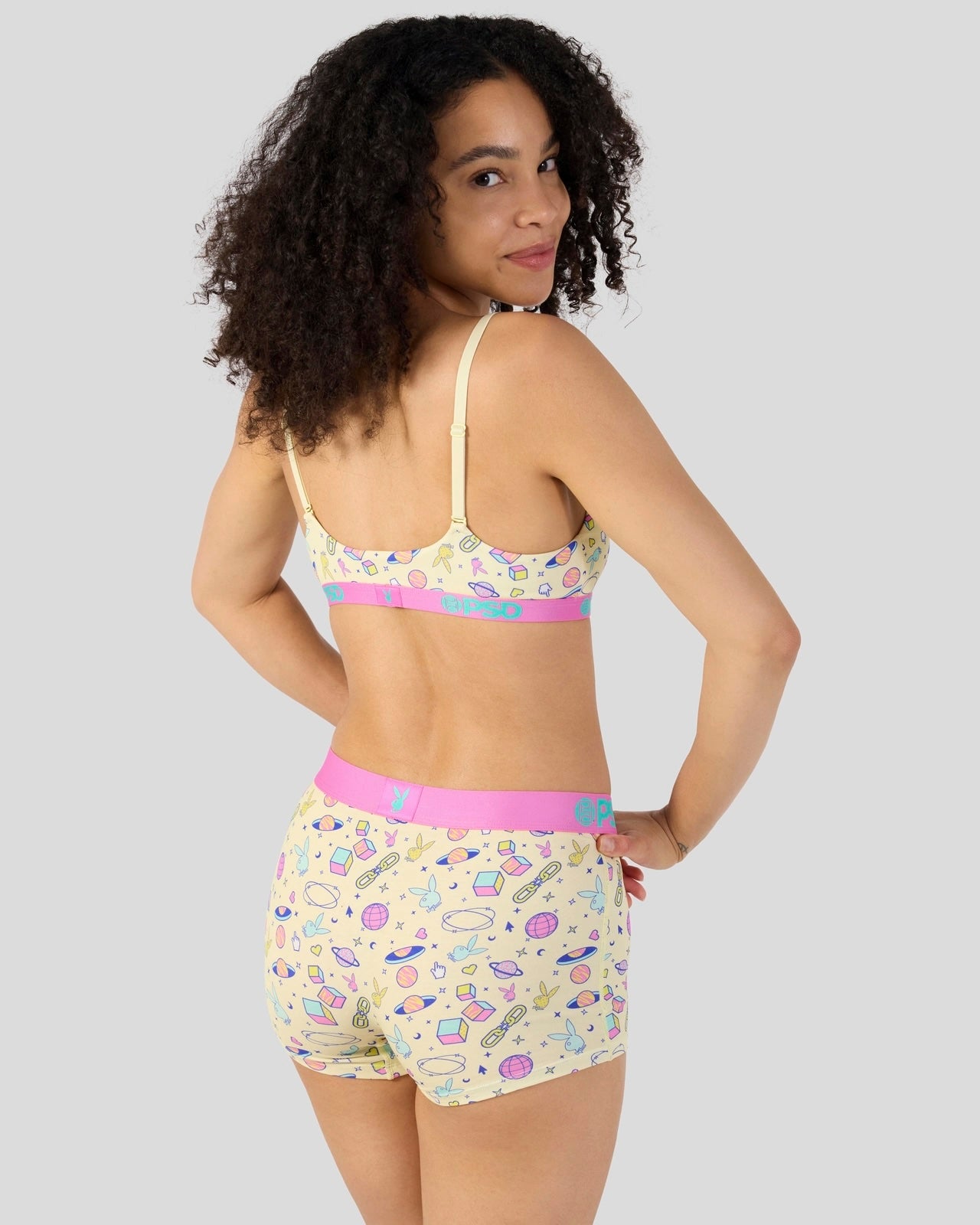 A woman modeling a matching two-piece lingerie set with a colorful candy print on a pale yellow background, featuring a pink waistband and a pink band on the bra.
