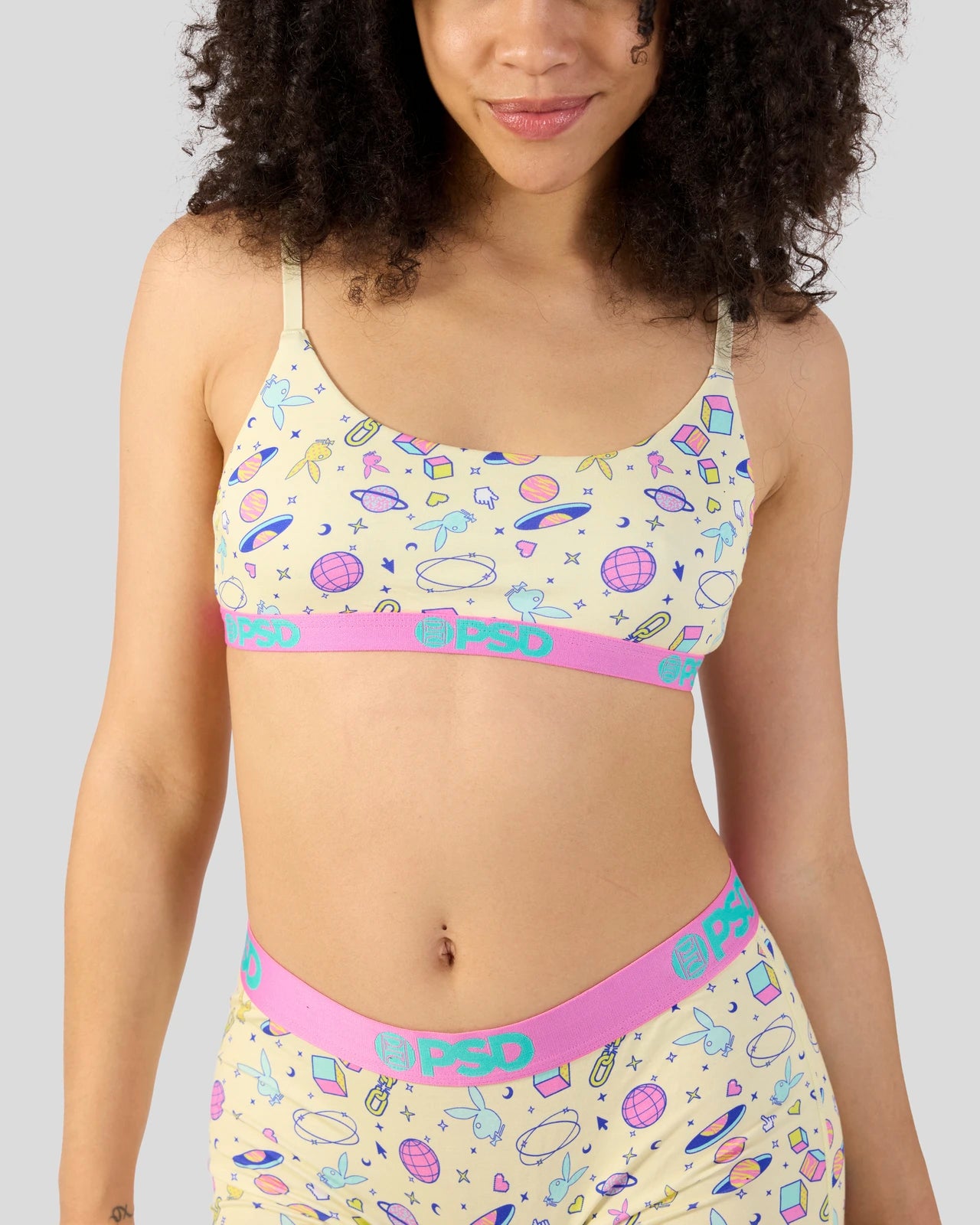 A woman wearing a matching two-piece pajama set with a pale yellow space-themed print and bright pink elastic waistbands. The set features a bra top and coordinating pants with the brand name 'PSD' printed on the waistbands.