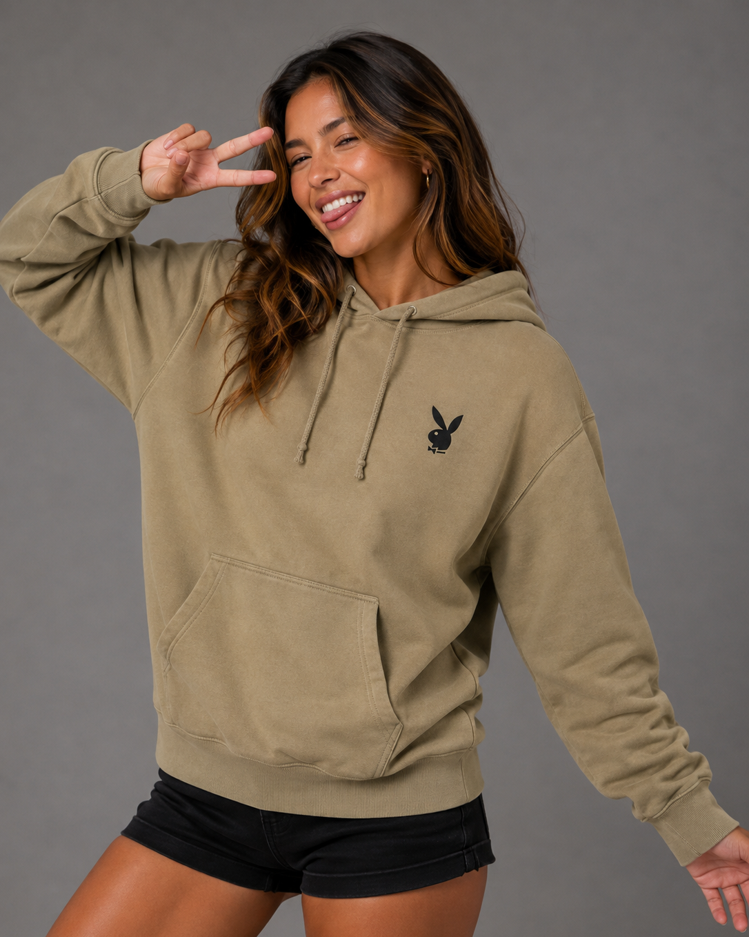 A woman modeling a khaki olive green pullover hoodie with a small black embroidered Playboy bunny logo on the left chest, paired with black shorts, against a solid gray studio background.