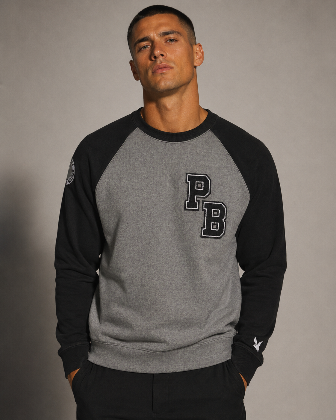 A man wearing a heather gray and black raglan sweatshirt with a large 'PB' graphic on the chest, posed against a neutral gray studio background.