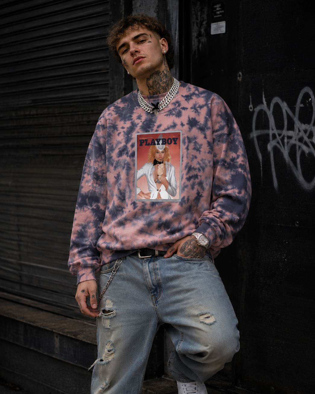A man wearing a pink and blue tie-dye sweatshirt with a vintage Playboy graphic on the front, paired with distressed light blue jeans and white sneakers, leaning against a dark graffiti-covered wall. #Streetwearmen