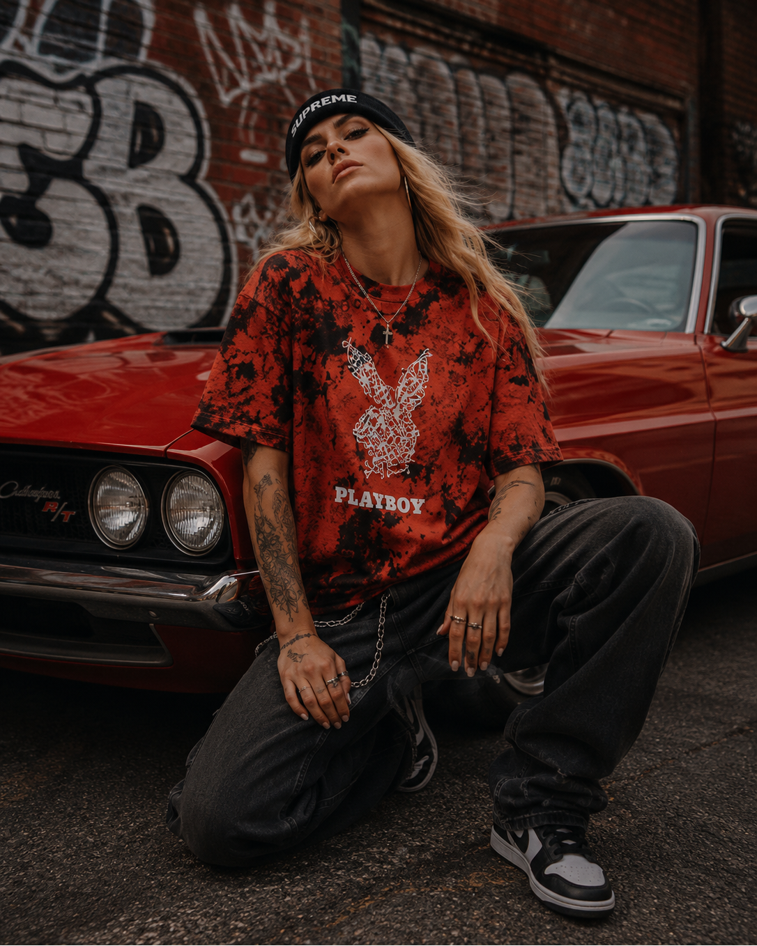 A woman kneels in front of a red classic car, wearing a red and black tie-dye Playboy t-shirt, black sweatpants, and a black beanie with the word 'PLAYBOY' on it. The background features a brick wall covered in graffiti. #Streetwearwomen