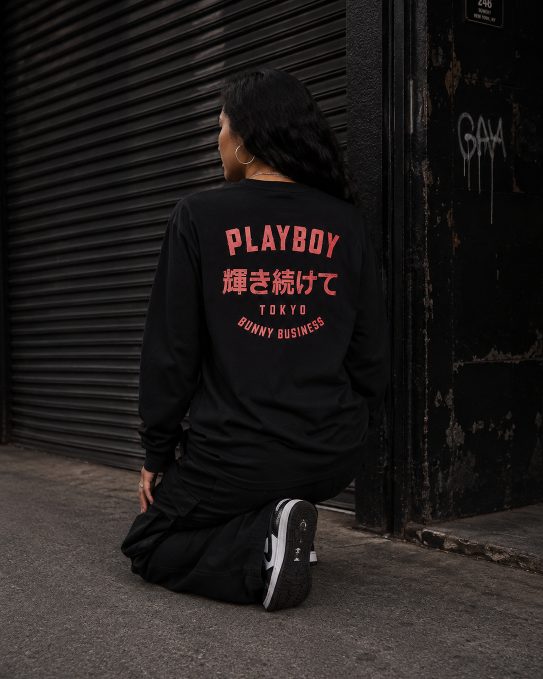 A person kneeling on a street in front of a closed metal garage door, wearing a black long-sleeve shirt from the back. The shirt features a large graphic print in coral pink and white on the back reading 'PLAYBOY', Japanese characters, 'TOKYO', and 'BUNNY BUSINESS'. #Streetwearwomen