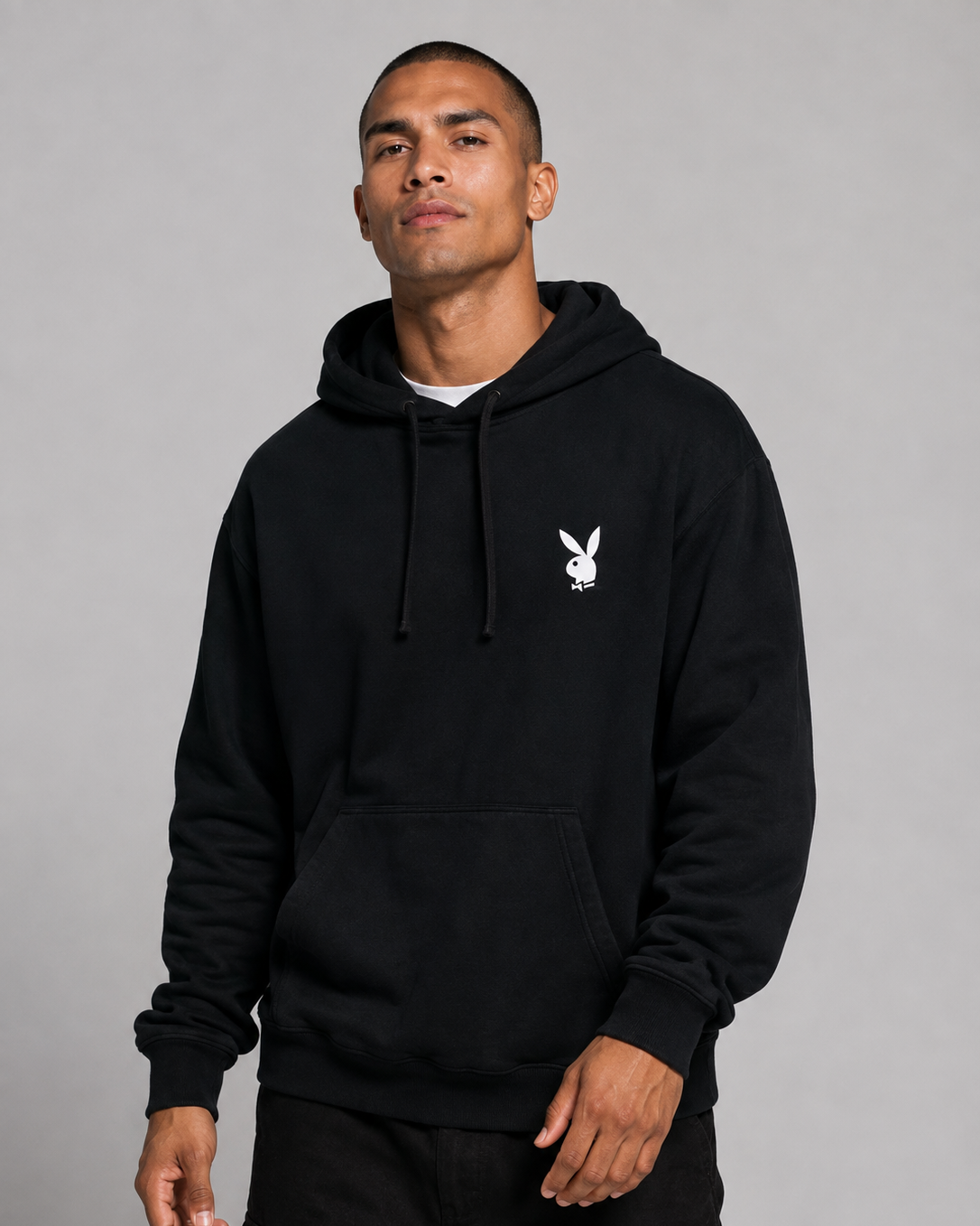 A man wearing a black pullover hoodie with a small white Playboy bunny logo on the left chest, photographed against a plain gray studio background.