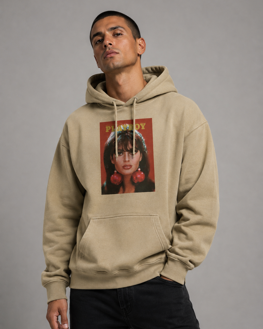 A man wearing a beige pullover hoodie with a vintage-style graphic print on the chest, paired with black pants, photographed against a neutral gray studio background.