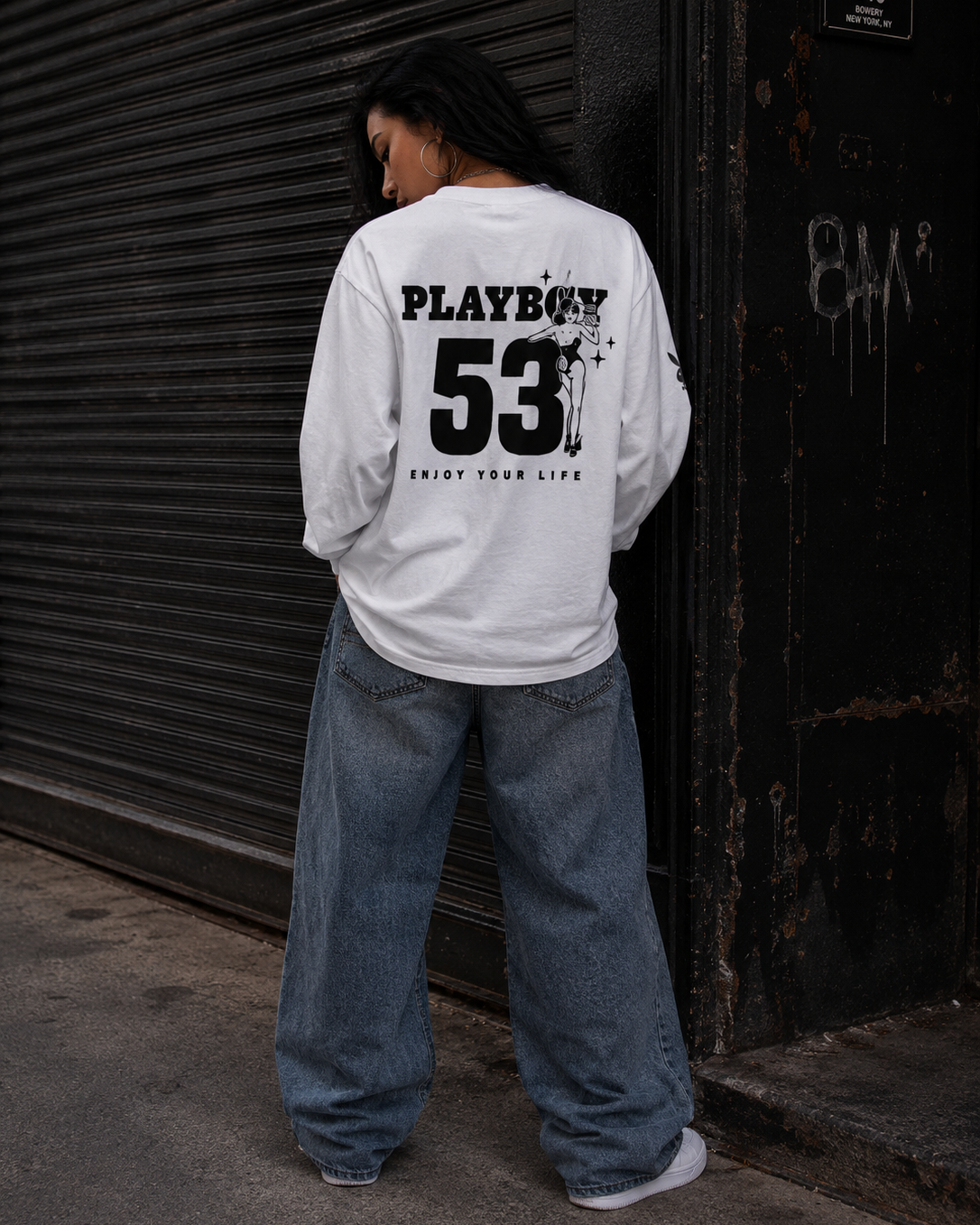 A person standing with their back to the camera wearing a white long-sleeve t-shirt with a large black 'PLAYBOY 53' graphic on the back, paired with wide-leg blue jeans. #Streetwearwomen
