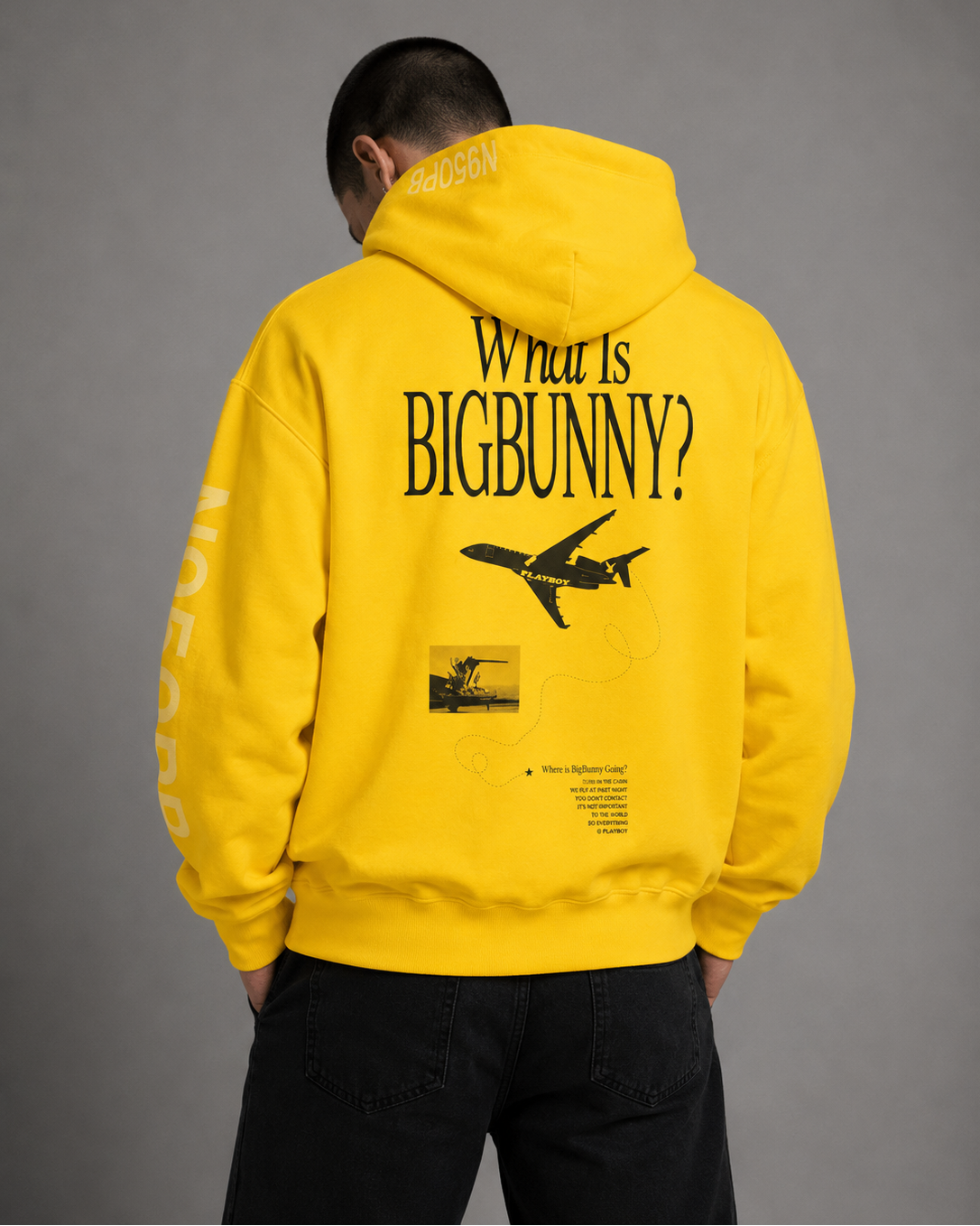 A person shown from the back wearing a bright yellow hoodie with large black text reading 'What Is BIGBUNNY?' and an airplane graphic on the back, paired with black pants against a plain gray studio background.