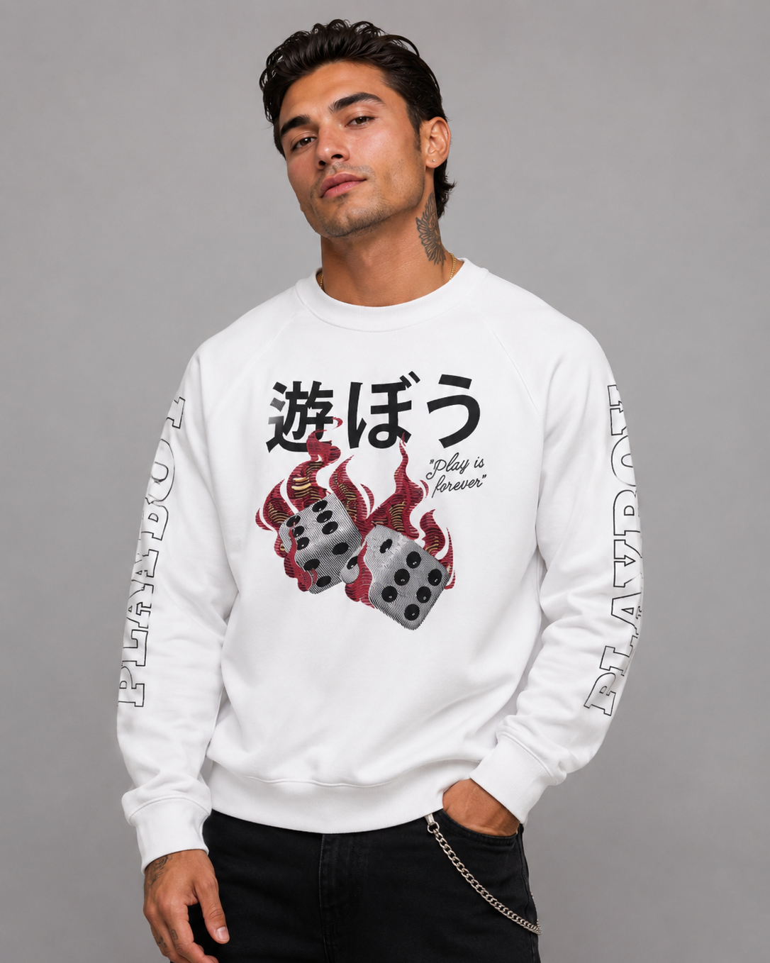 A man wearing a white crewneck sweatshirt with a graphic of two red and gray dice with flames, accompanied by Japanese text and 'Play is forever' on the front, and 'PLAY DOUGH' printed down both sleeves. He is wearing black pants against a solid gray studio background.