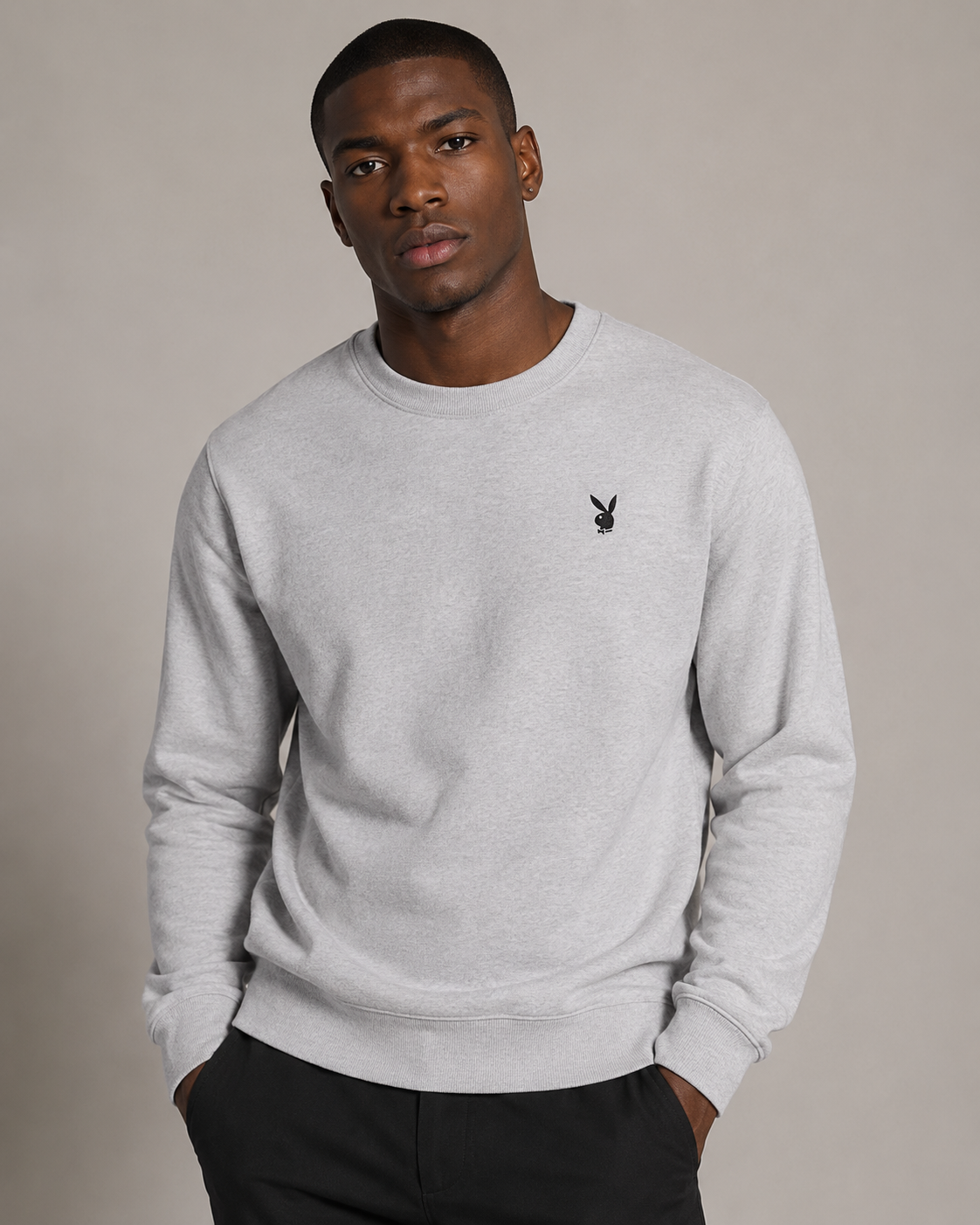 A man wearing a light gray heather crewneck sweatshirt with a small black embroidered Playboy bunny logo on the left chest, paired with black pants, against a solid beige studio background.