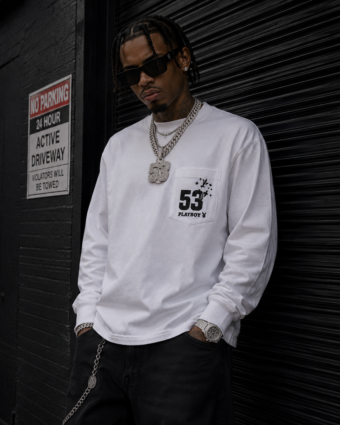 A man wearing a white long-sleeve crewneck shirt with a black graphic print on the pocket, styled with black pants, gold chains, and sunglasses, standing in front of a black metal roll-up door. #Streetwearmen
