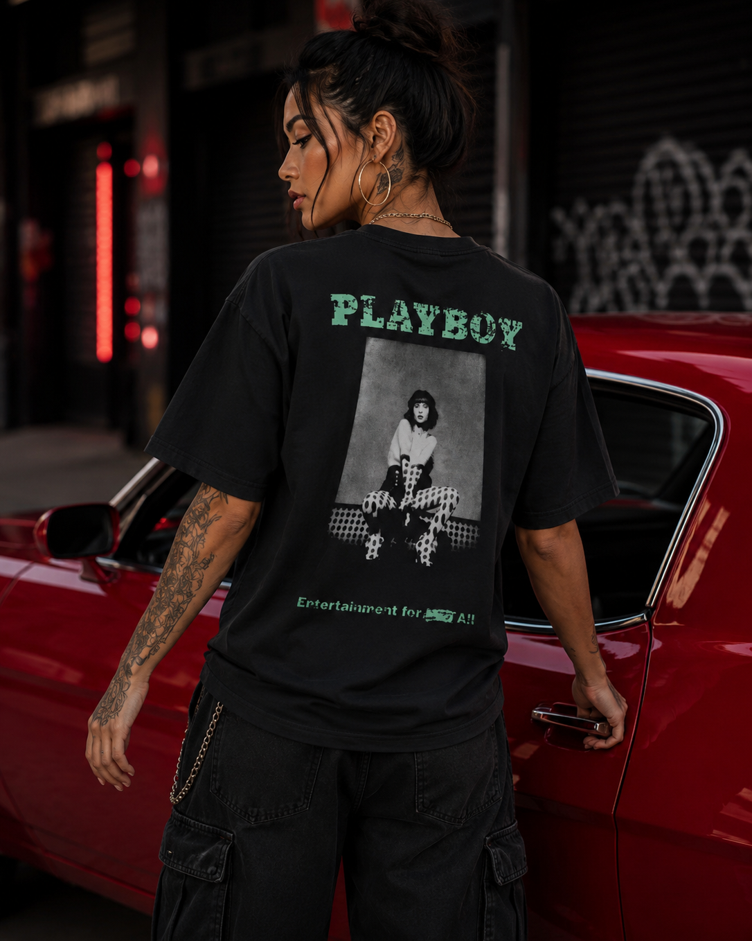 A woman shown from the back wearing an oversized black t-shirt with a green PLAYBOY graphic and photo print, black cargo pants, standing beside a red muscle car on a city street at night. #Streetwearwomen