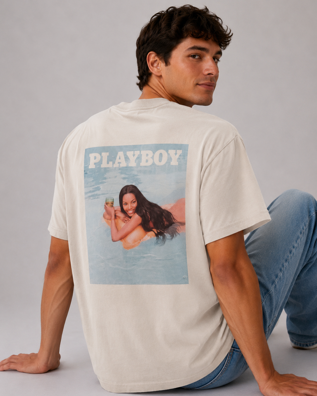 A man wearing a cream-colored Playboy t-shirt with a large back graphic print, paired with blue jeans, sitting against a neutral gray studio background.