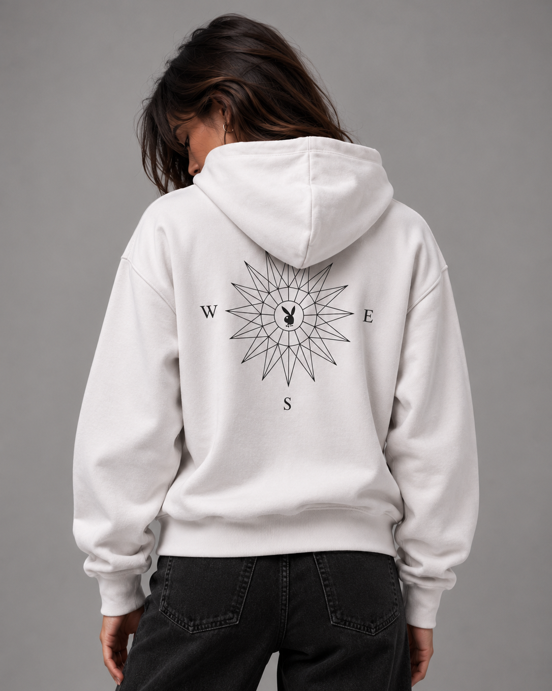 A person shown from the back wearing a white pullover hoodie with a large black compass rose graphic and the letters W, E, S on the back, paired with dark jeans against a plain gray studio background.