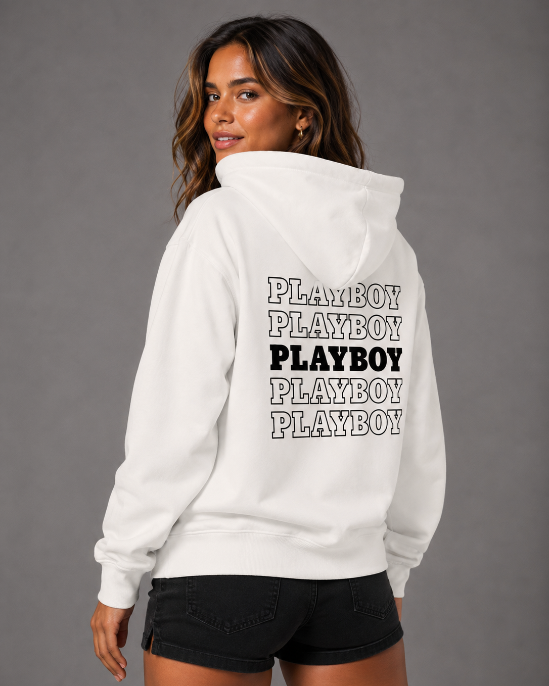 A woman shown from the back wearing a white hoodie with 'PLAYBOY' printed five times in black on the back, paired with black denim shorts, against a solid gray studio background.