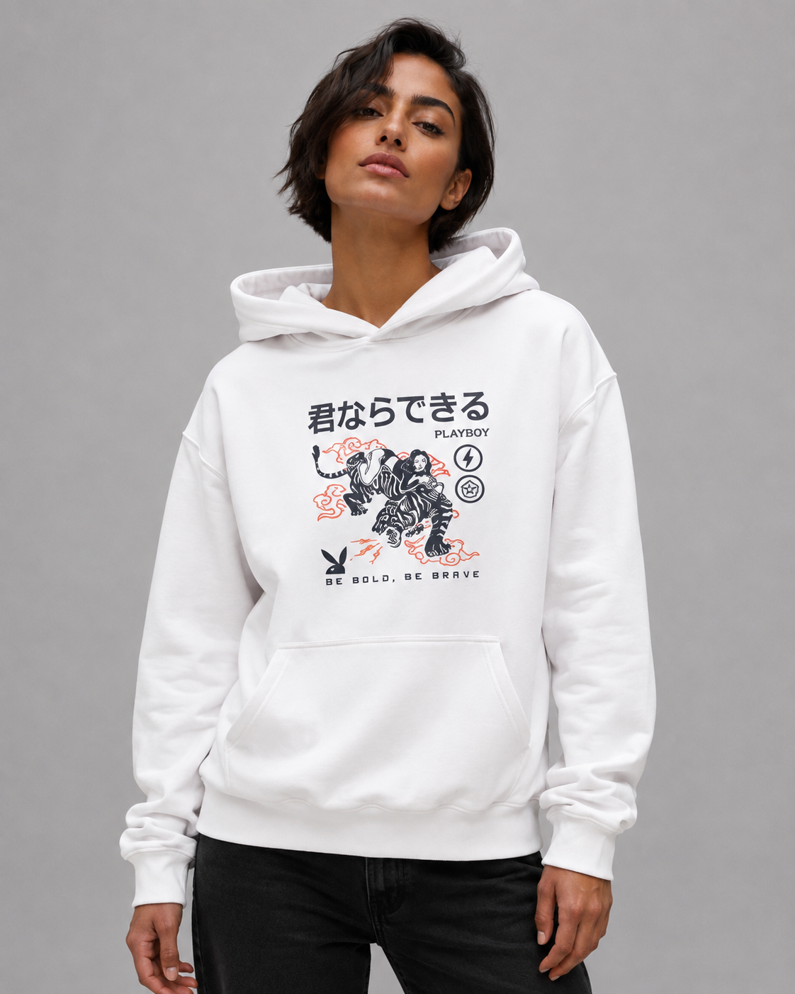 A woman with short dark hair wearing a white Playboy Be Bold hoodie with a tiger graphic and Japanese text, against a solid gray studio background. #Streetwearwomen