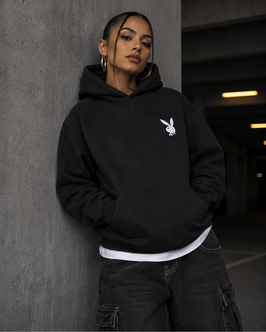A woman wearing a black pullover hoodie with a white Playboy bunny logo on the chest, posing against a concrete pillar in a dimly lit parking structure. #Streetwearwomen