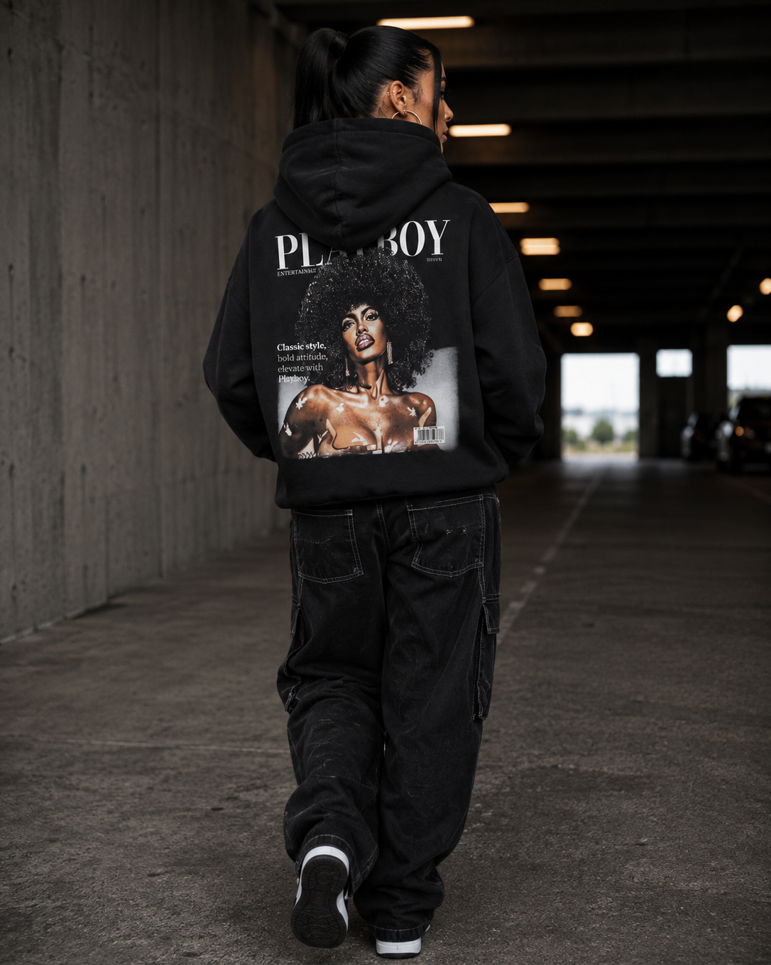 A person walking away in a concrete tunnel wearing a black hoodie with a large Playboy magazine cover graphic printed on the back, paired with black cargo pants and black sneakers. #Streetwearwomen