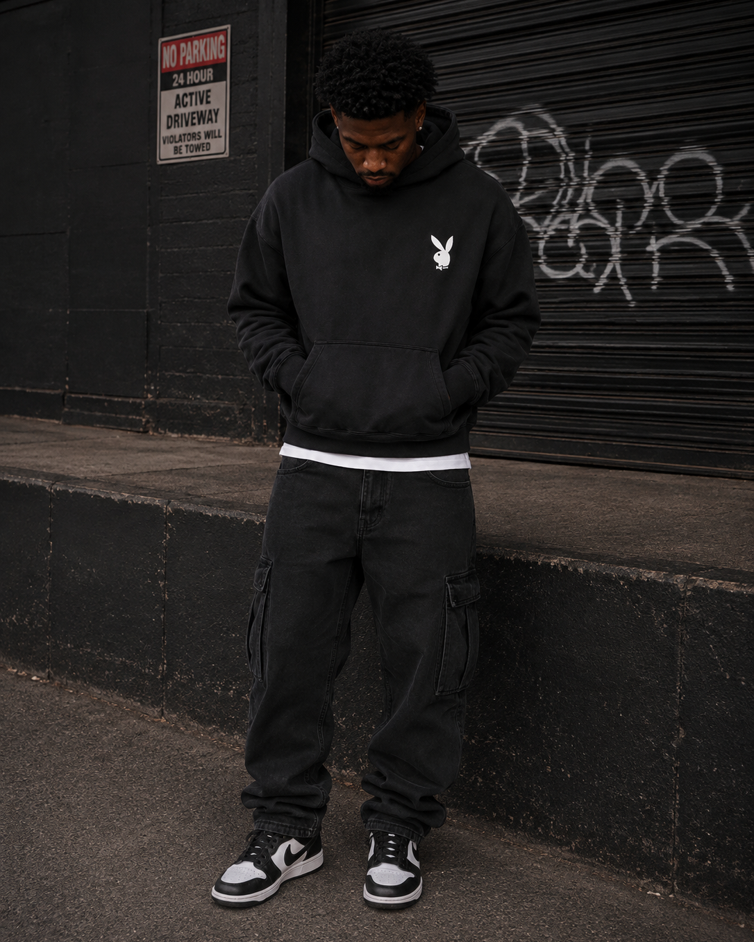 A man wearing a black hoodie with a small white Playboy bunny logo on the left chest, black cargo pants, and black-and-white sneakers, standing in front of a dark metal shutter with graffiti. #Streetwearmen