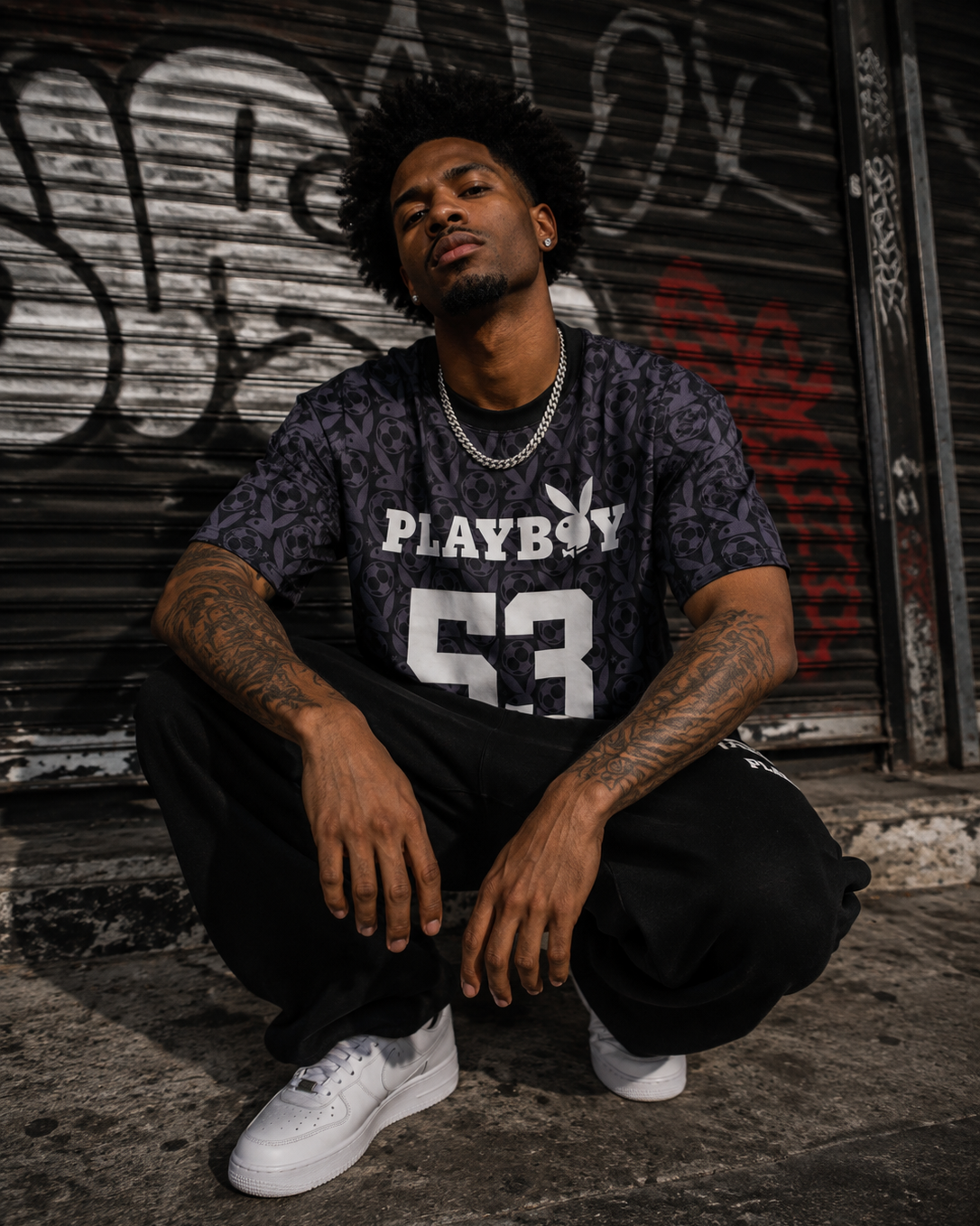A man crouches in front of a graffiti-covered metal shutter wearing a dark washed t-shirt with a 'PLAYBOY 53' print, paired with white sneakers. #Streetwearmen
