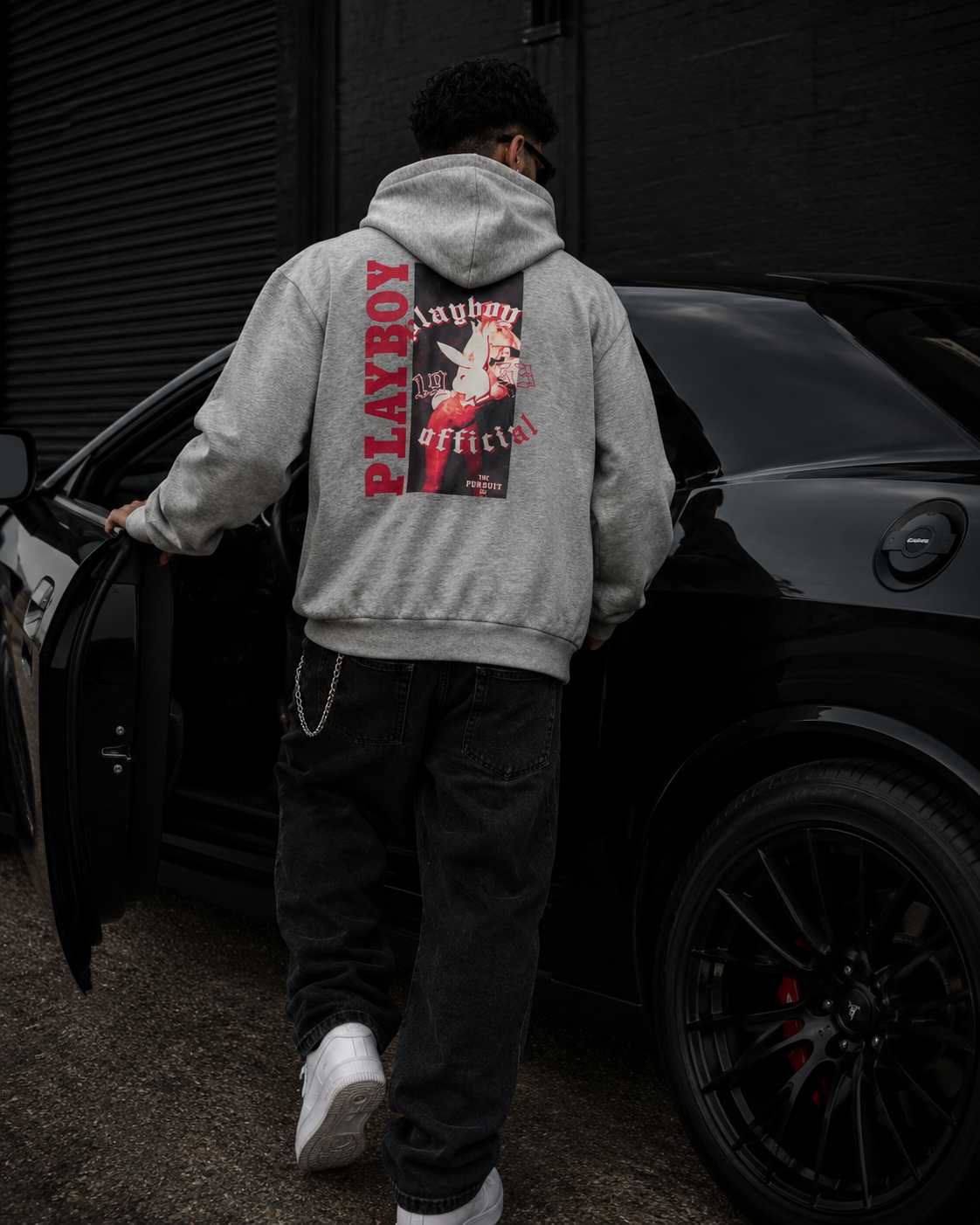 A man viewed from behind wearing a light gray Playboy hoodie with a large red and black graphic on the back, standing next to an open door of a black car. #Streetwearmen