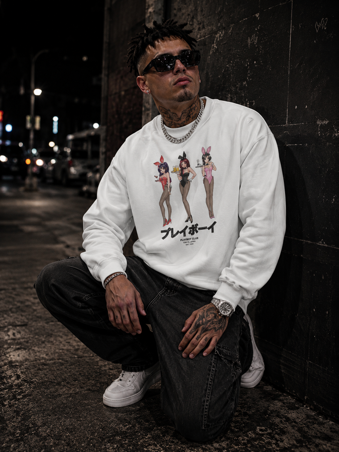 A man wearing a white crewneck sweatshirt with a graphic of three pin-up girls and Japanese text, paired with black jeans and white sneakers, crouching against a dark wall at night. #Streetwearmen