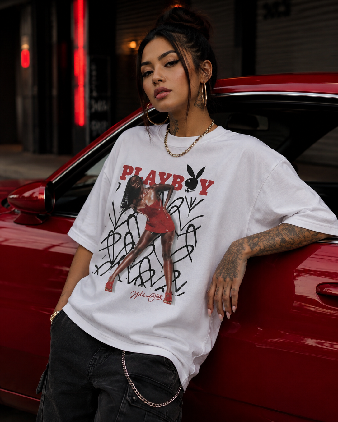 A woman leans against a red car wearing an oversized white graphic T-shirt with a red Playboy logo and black graffiti-style text, paired with dark cargo pants. #Streetwearwomen