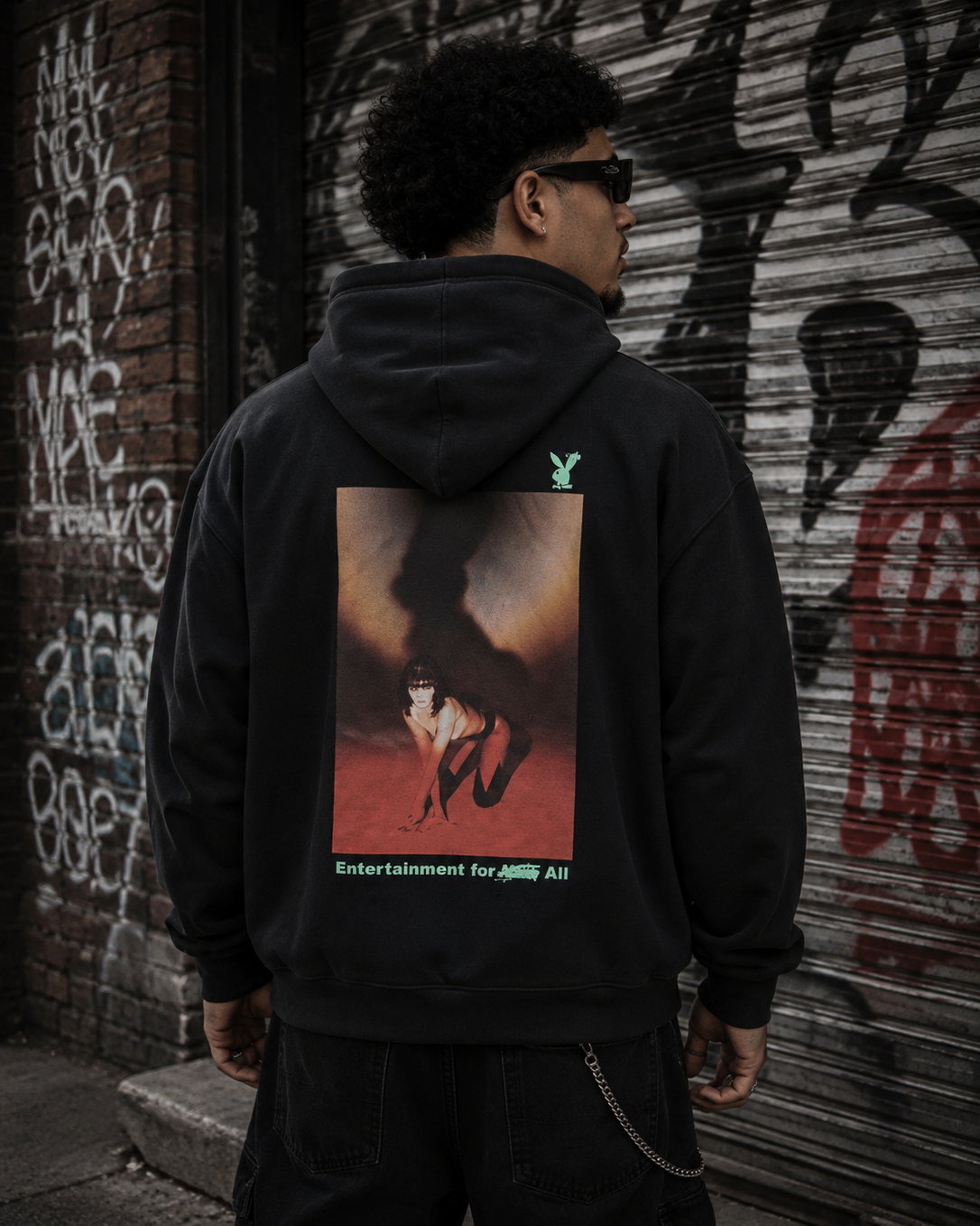 A man wearing a black hoodie with a large graphic print on the back, photographed from behind in an urban setting with a graffiti-covered metal shutter background. #Streetwearmen