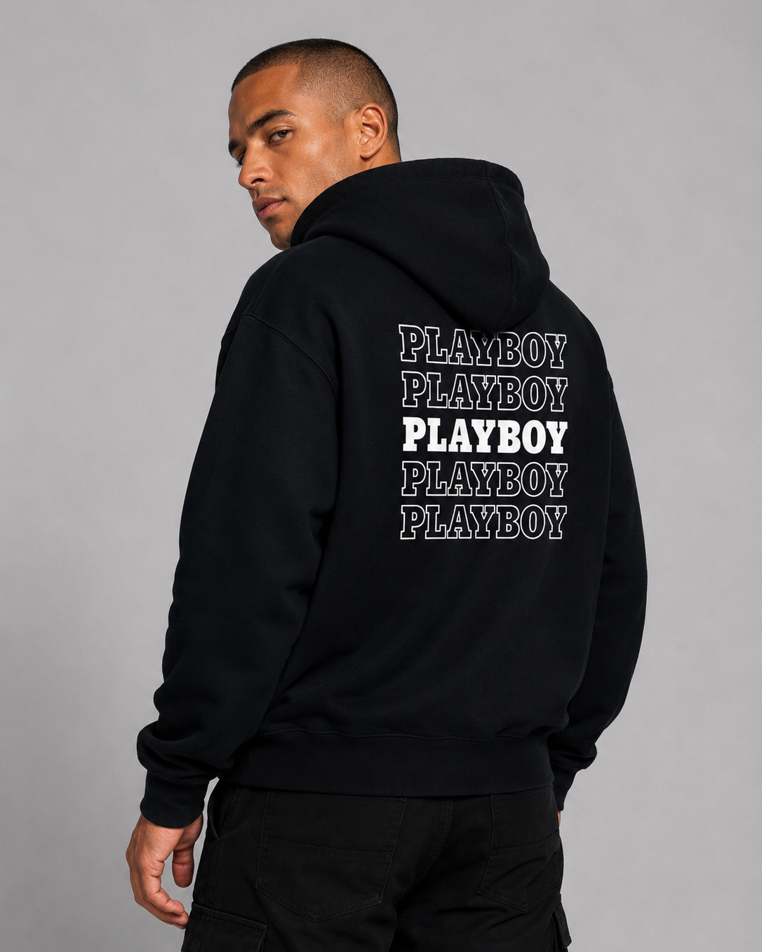 A man wearing a black hoodie from the back, featuring the brand name 'PLAYBOY' printed five times in white and gray block letters on the back, set against a plain gray studio background.