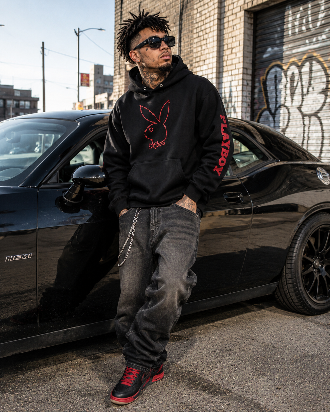 A man wearing a black Playboy hoodie with a red bunny logo and 'PLAYBOY' text on the sleeve, paired with dark gray jeans and black sneakers, standing next to a black car in an urban setting. #Streetwearmen