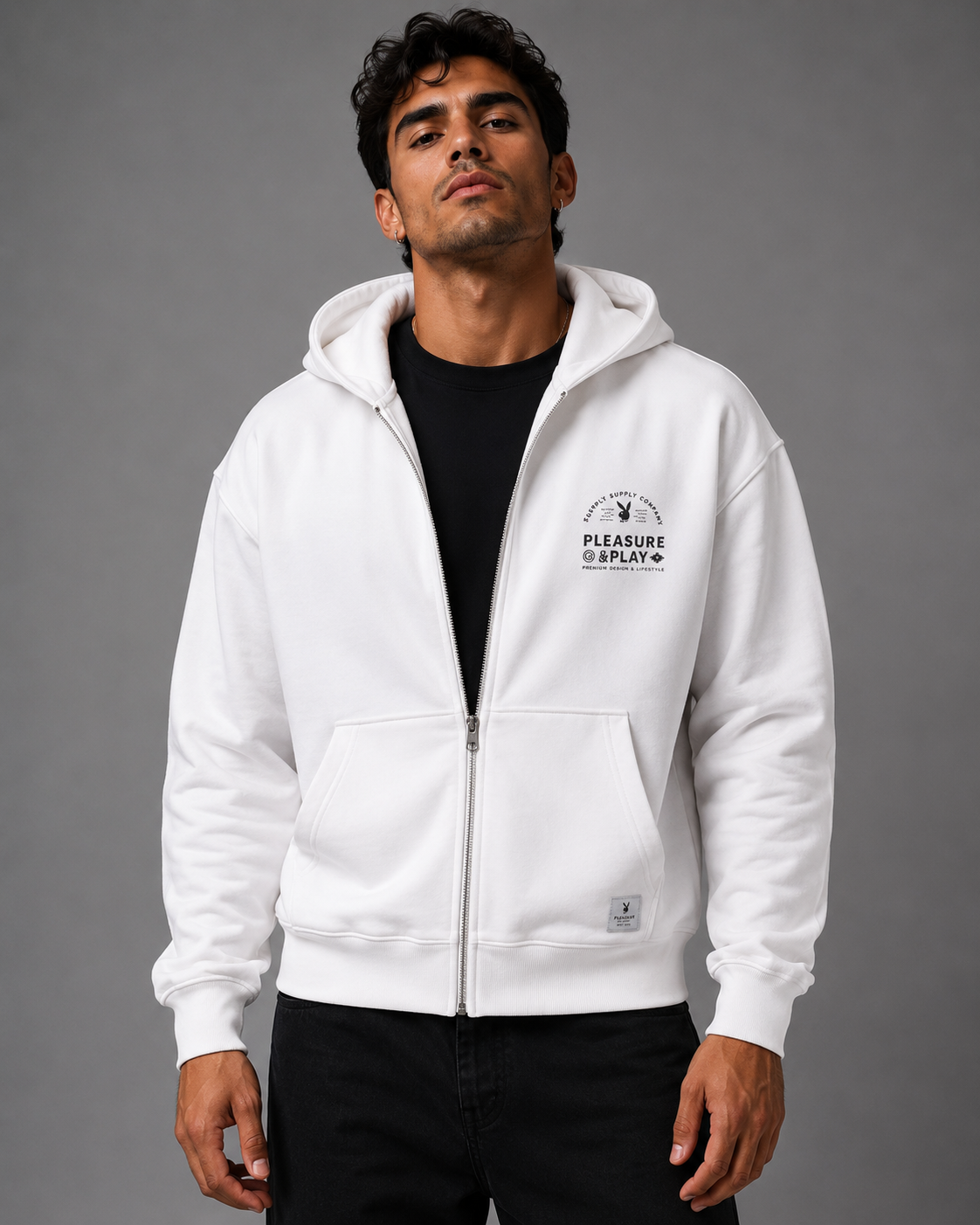 A man wearing a white zip-up hoodie with a black graphic print on the left chest, layered over a black t-shirt and black pants, photographed against a solid gray studio background.