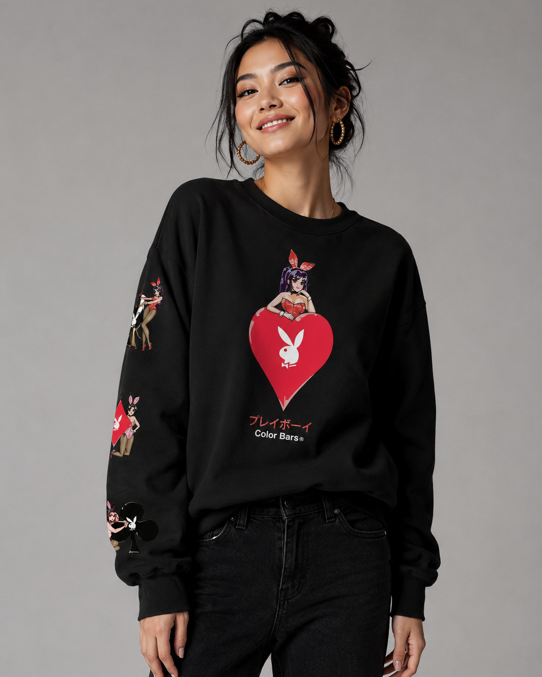 A woman modeling a black crewneck sweatshirt featuring a large red heart graphic with the Playboy bunny logo on the front, along with rabbit-themed illustrations on the sleeves. She is wearing black jeans against a solid gray studio background.