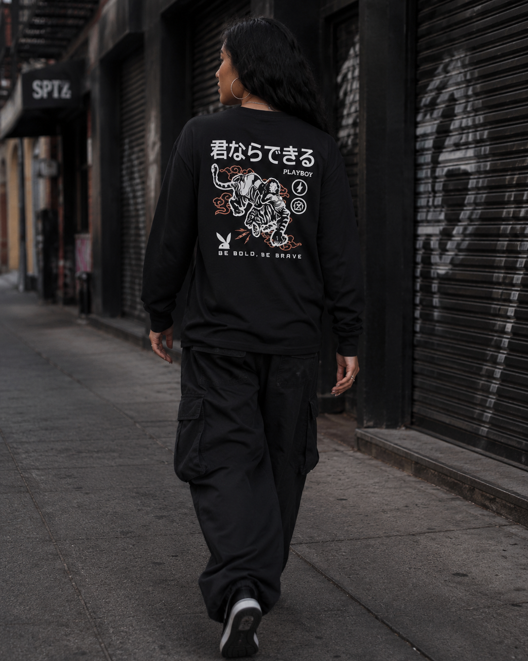 A person walking down an urban street away from the camera, wearing a black long-sleeve shirt with a large white and red graphic print on the back, paired with black cargo pants. #Streetwearwomen
