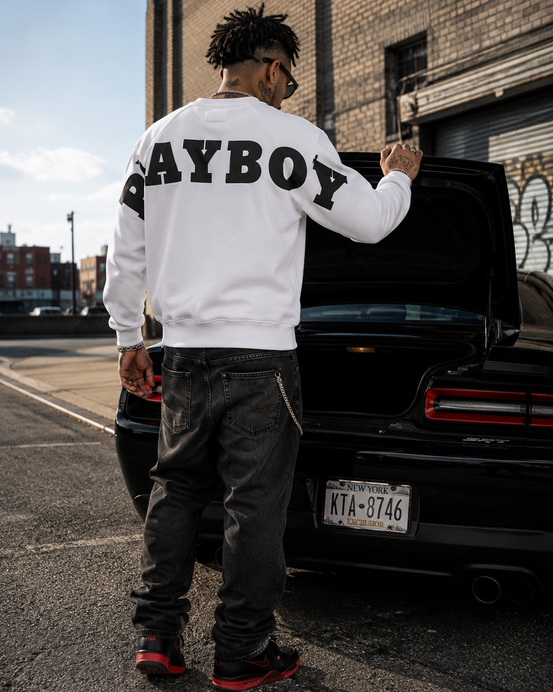 A man stands with his back to the camera, wearing a white long-sleeve sweatshirt with 'PAYBOY' printed in black across the back, paired with dark gray jeans and black sneakers with red accents. He is standing next to the open trunk of a black car in an urban street setting. #Streetwearmen