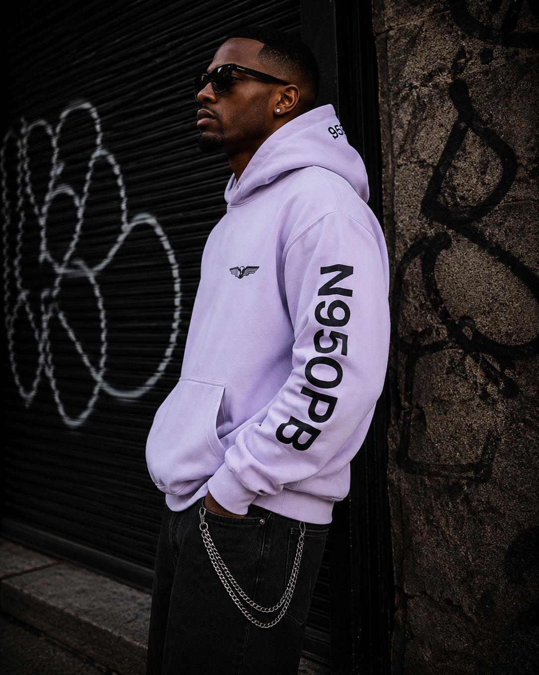 A man wearing a light purple/lavender hoodie with black text 'N950PB' printed down the right sleeve and a small wing logo on the left chest, standing against a graffiti-covered dark metal shutter. #Streetwearmen