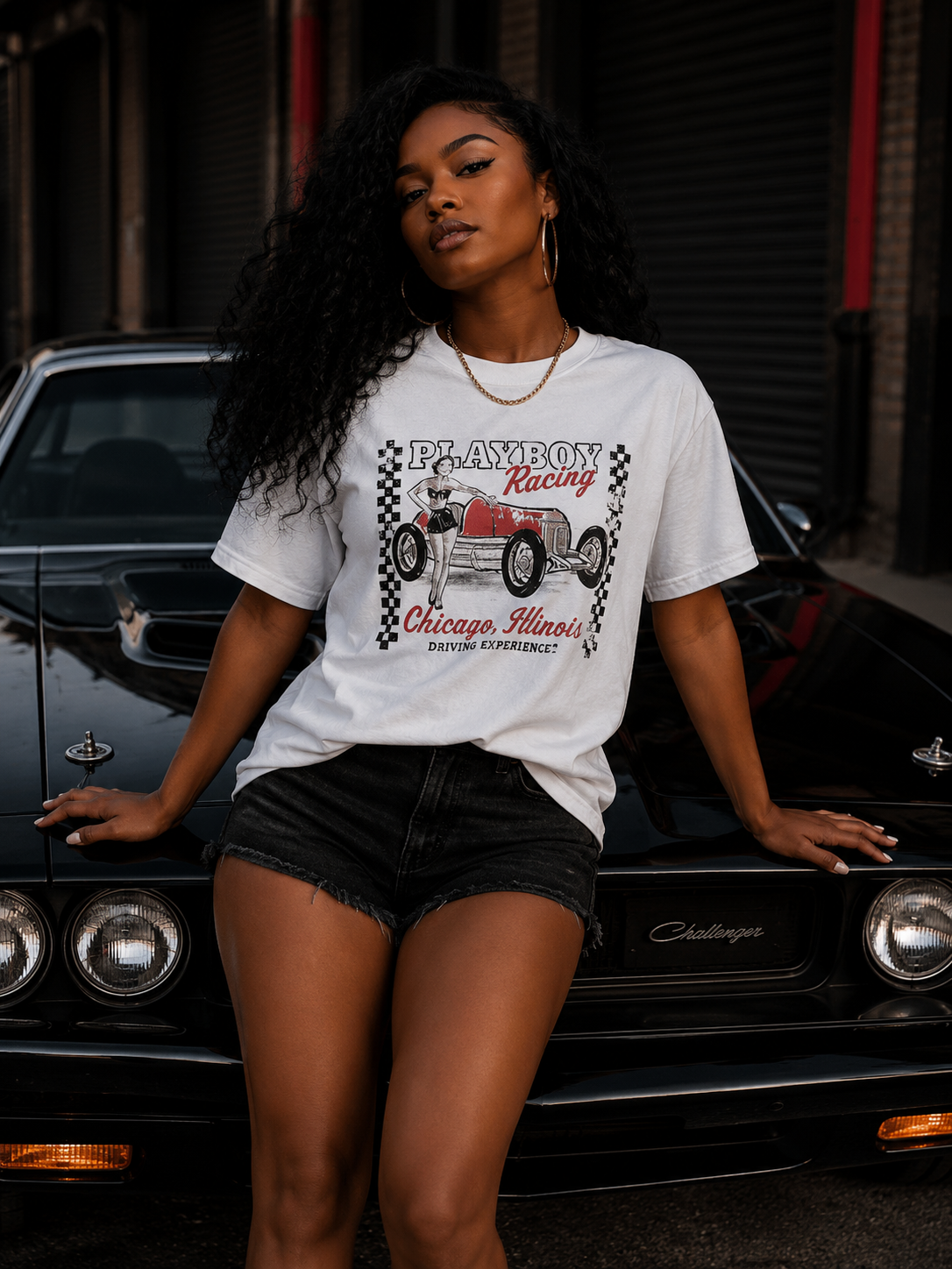 A woman posing in front of a black classic car, wearing a white graphic t-shirt that reads 'Blayboy Racing Chicago, Illinois' with a vintage hot air balloon design, paired with black denim shorts. #Streetwearwomen