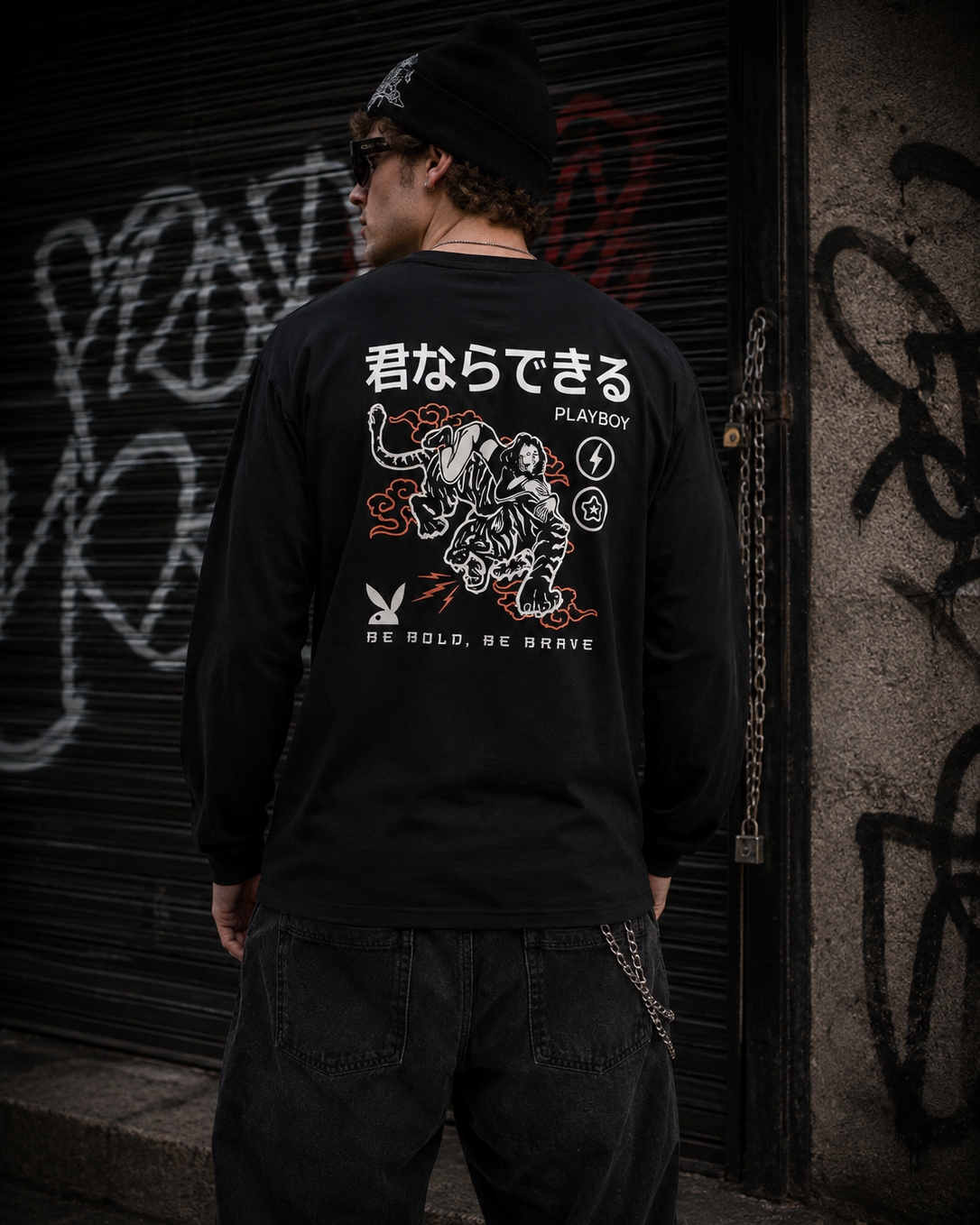 A man viewed from behind wearing a black long-sleeve PLAYBOY shirt with a large graphic print on the back, standing in front of a graffiti-covered metal shutter. #Streetwearmen