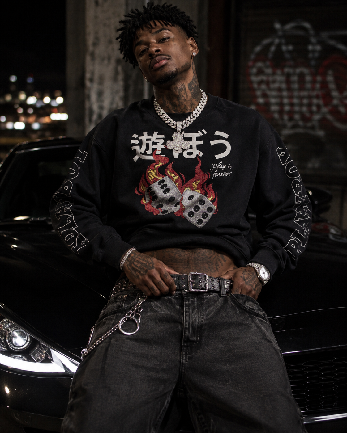 A man wearing a black crewneck sweatshirt with a graphic of two dice and Japanese characters, paired with dark gray jeans and a silver chain necklace, posing in front of a black car at night. #Streetwearmen