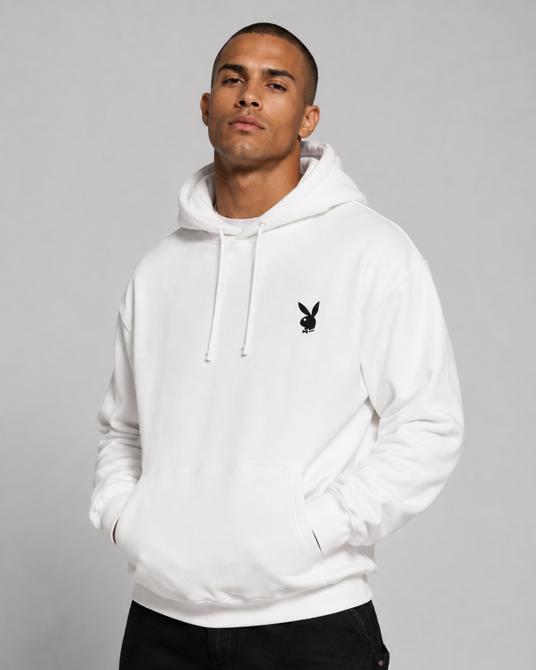 A man wearing a white pullover hoodie with a small black Playboy bunny logo on the left chest, paired with black pants, against a solid gray studio background.