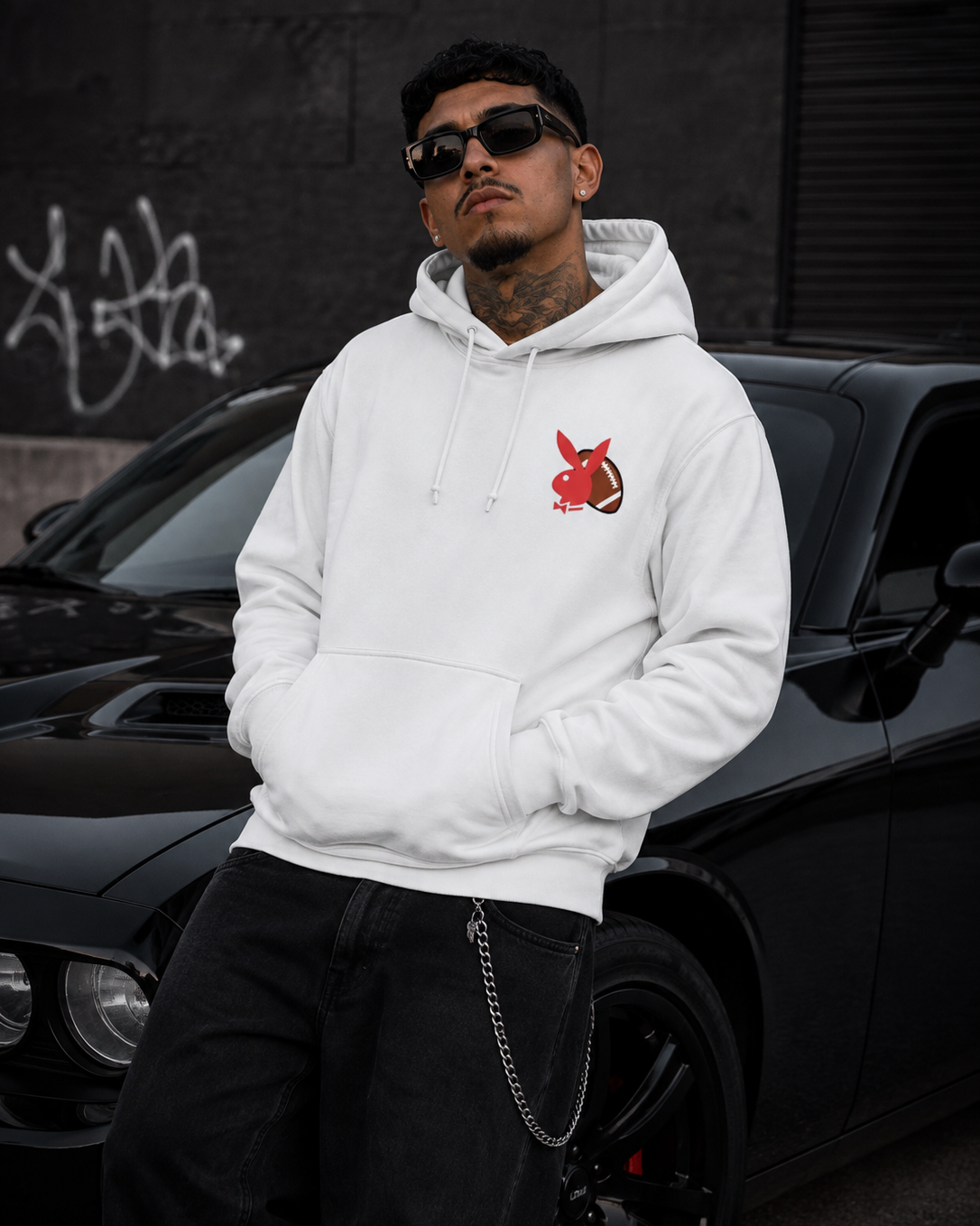 A man wearing a white pullover hoodie with a red rabbit graphic on the chest, black pants, and sunglasses, standing in front of a black car. #Streetwearmen