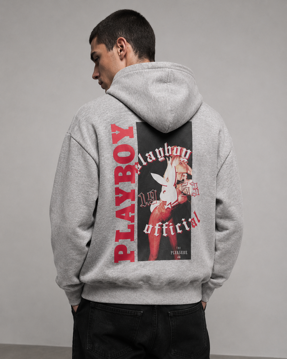 A man wearing a light gray heathered hoodie from the back, featuring a large graphic print with red 'PLAYBOY' lettering and a black-and-white bunny logo. He is also wearing black jeans against a neutral gray studio background.