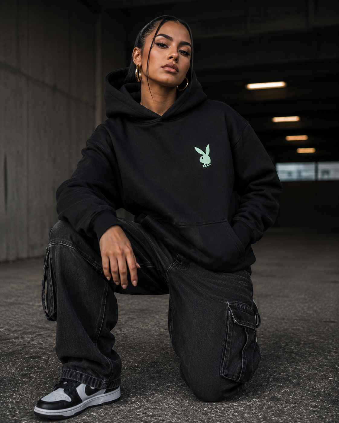 A model wearing a black hoodie with a small mint green Playboy bunny logo on the left chest, paired with black cargo pants and sneakers, posing in a concrete tunnel. #Streetwearwomen