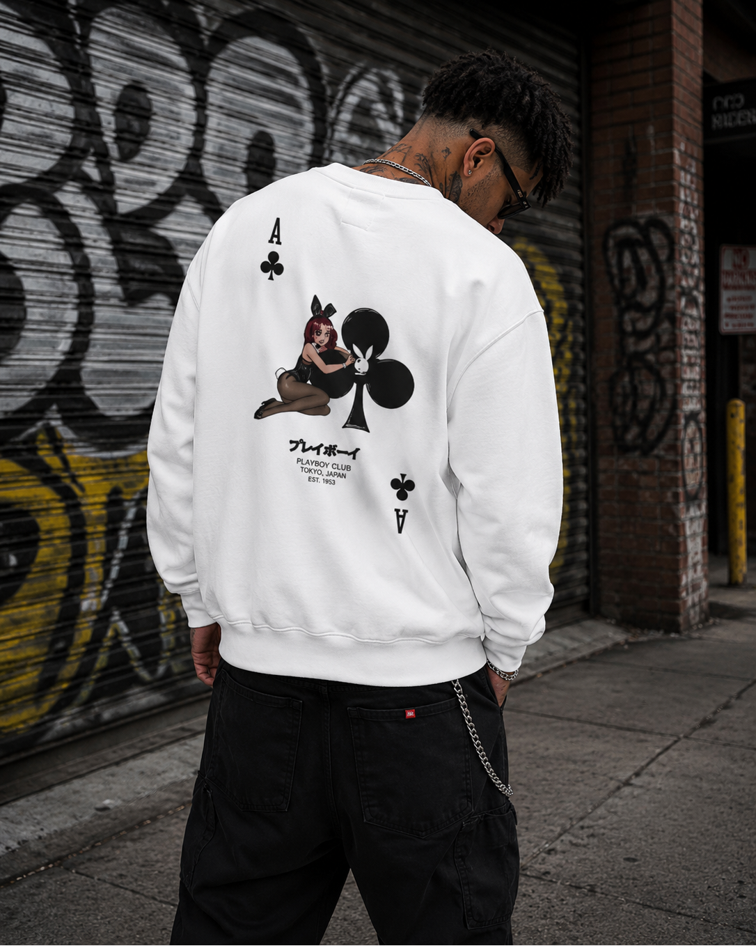 A man stands with his back to the camera wearing a white crewneck sweatshirt with an ace of clubs playing card graphic on the back, in front of a graffiti-covered urban background. #Streetwearmen