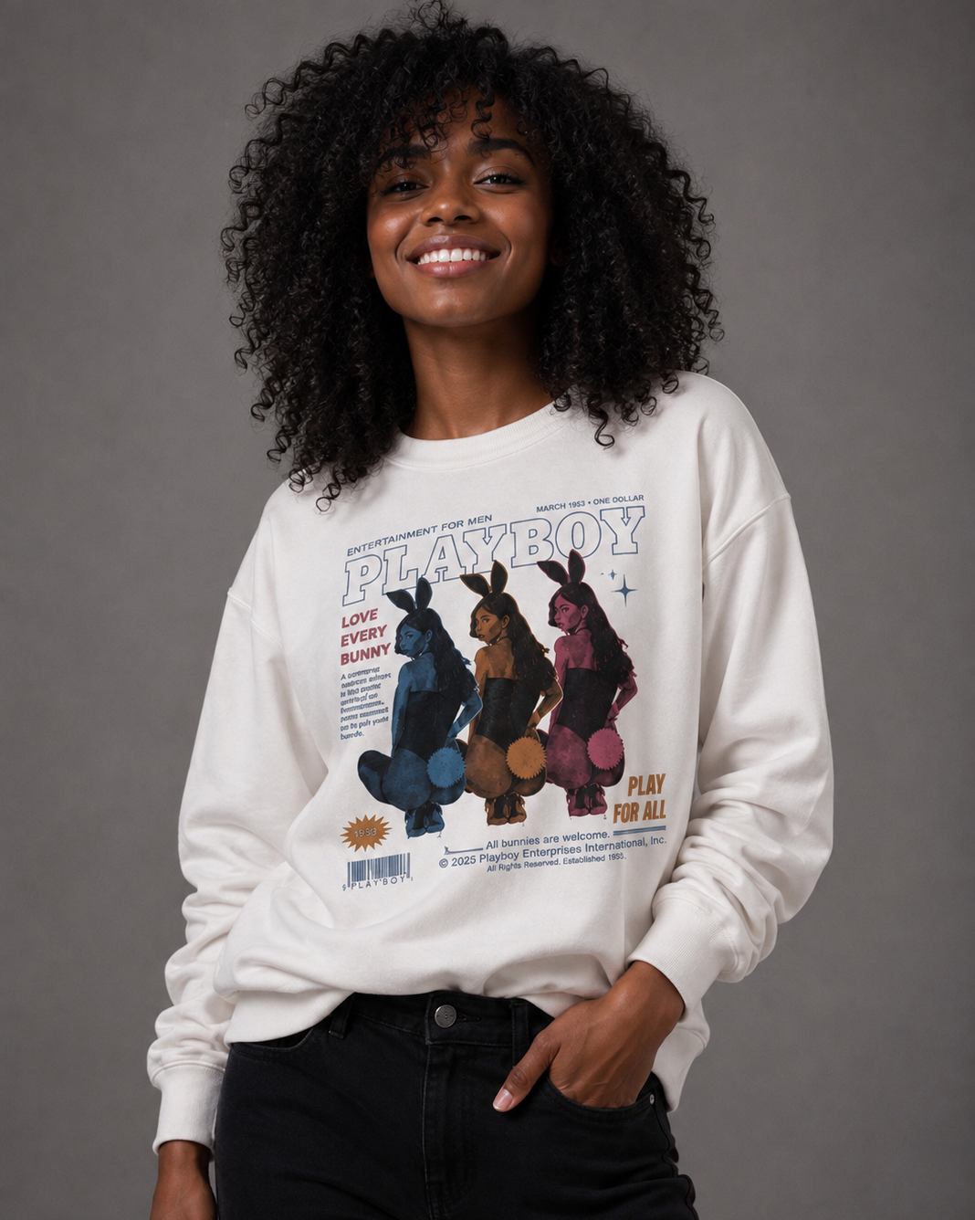 A woman with curly black hair wearing a white Playboy sweatshirt with a vintage-style bunny graphic and black jeans, standing against a solid gray studio background.